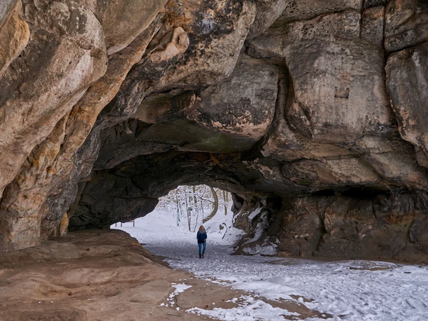 Rock Gate Cowshed in Winter Person in blue jacket walks through a large, natural rock arch of the cowshed; the ground is covered with snow, trees in the background.