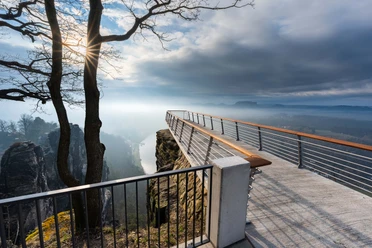Uitkijkplatform met leuningen boven een kloof, op de achtergrond een mistig landschap en rotsen; de zon schijnt door kale boomtakken.