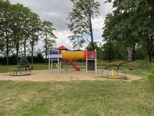 Spielplatz im Erlebnisbad Rathewalde Spielplatz mit roter Rutsche, gelbem Tunnel, Wippe und Karussell auf Sandfläche, umgeben von Bäumen und Wiese unter bewölktem Himmel.