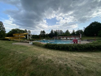 Buitenzwembad Rathewalde Een buitenzwembad met een gele waterglijbaan, een groot bad en een rode olifantenglijbaan, omringd door gras en bomen onder een bewolkte hemel.