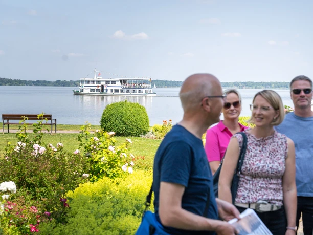 weisse flotte.jpg Ein Ausflugsschiff der Weißen Flotte fährt über einen ruhigen See. Im Vordergrund genießen Menschen einen Spaziergang am Ufer.