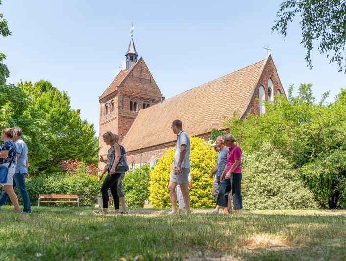 sehenswürdigkeiten.jpg Vor einem Backsteingebäude ragt der Kirchturm auf, während Menschen auf dem grünen Rasen spazieren.