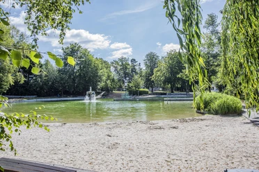 Waldbad 1 Kleiner See mit Sandstrand, umgeben von grünen Bäumen und einem Springbrunnen in der Mitte.