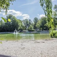 Waldbad 1 Kleiner See mit Sandstrand, umgeben von grünen Bäumen und einem Springbrunnen in der Mitte.