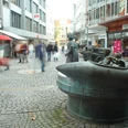 Passanten flanieren an einer Brunnen-Skulptur auf einer belebten Einkaufsstraße entlang.
