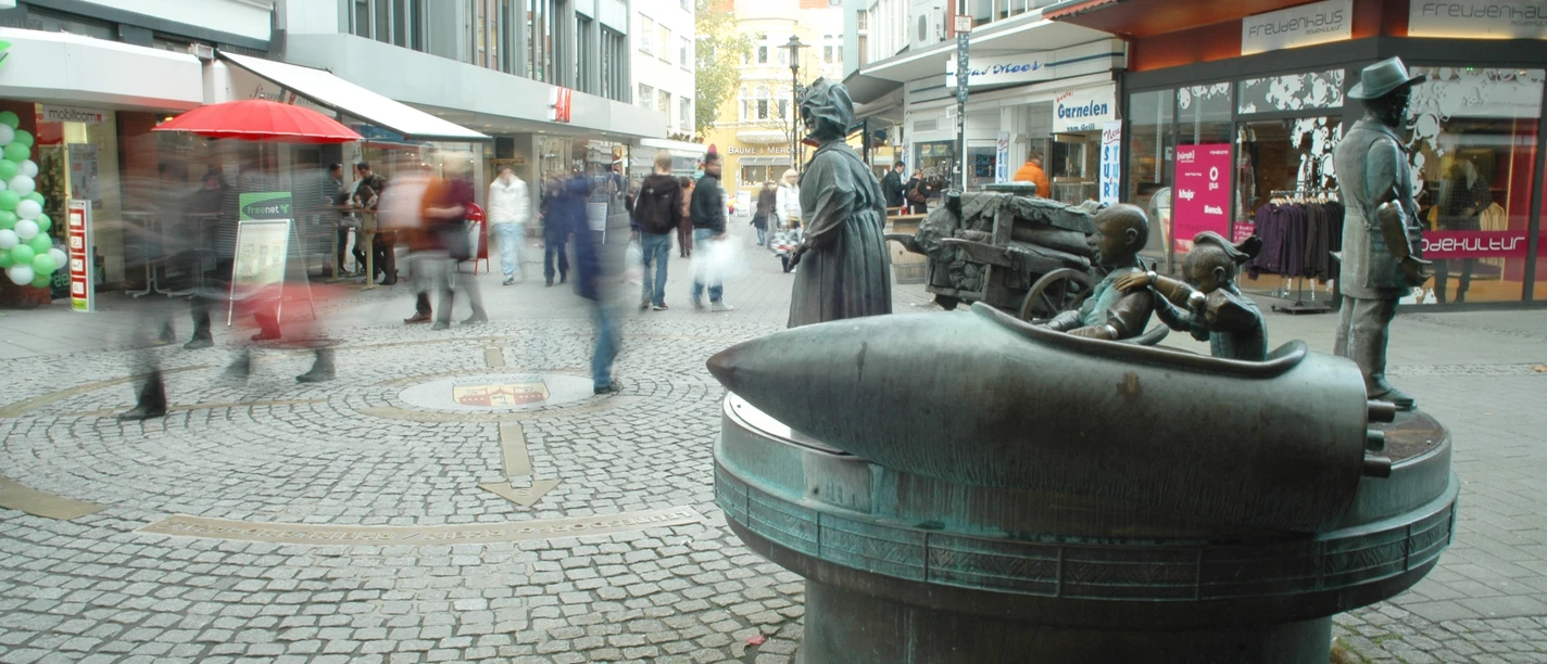 Passanten flanieren an einer Brunnen-Skulptur auf einer belebten Einkaufsstraße entlang.