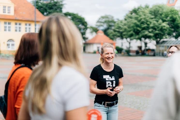 Kiez-Tour Bielefeld.jpg Lächelnde Frau in schwarzem T-Shirt steht in einer kleinen Gruppe auf einem gepflasterten Platz.