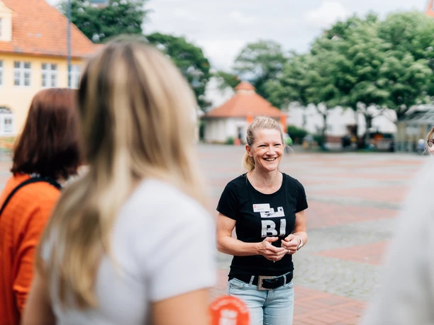 Kiez-Tour Bielefeld.jpg Lächelnde Frau in schwarzem T-Shirt steht in einer kleinen Gruppe auf einem gepflasterten Platz.