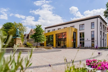 Gemeindebibliothek Steinhagen (C) pro Wirtschaft GT, Mario Wallenfang.jpeg Ein modernes Gebäude mit goldener Glasfront und einer Statue auf einem gepflasterten Platz bei Sonnenschein.