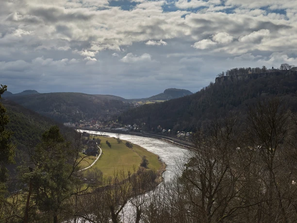 River landscape with wooded hills, a fortress on a hill to the right and a village on the riverbank under a cloudy sky.