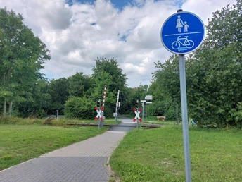 Bahnübergang Braker Weg Fußgänger- und Radweg am Bahnübergang mit Schranken, umgeben von Bäumen und blauem Hinweiszeichen.