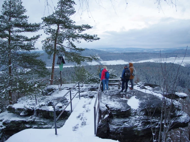Aussicht vom Signalstein Drei Personen in Winterkleidung stehen auf einem verschneiten Felsvorsprung mit Blick auf eine bewaldete, schneebedeckte Landschaft.
