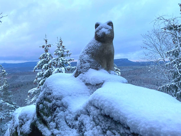 Stenen kattenbeeld op een besneeuwde rots, omringd door besneeuwde bomen en een brede, beboste vallei op de achtergrond.