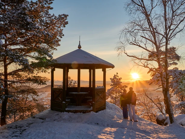 Een paviljoen in een besneeuwd bos bij zonsondergang, twee mensen staan naast elkaar en kijken naar de ondergaande zon.