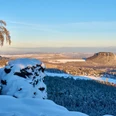 Aussicht vom Gohrisch Verschneite Felsen und ein kahler Baum im Vordergrund, mit Blick auf eine winterliche, sonnenbeschienene Hügellandschaft unter klarem, blauem Himmel.Snow-covered rocks and a bare tree in the foreground, with a view of a wintery, sunlit hilly landscape under a clear blue sky.Zasněžené skály a holý strom v popředí s výhledem na zimní, sluncem ozářenou kopcovitou krajinu pod jasně modrou oblohou.Pokryte śniegiem skały i nagie drzewo na pierwszym planie, z widokiem na zimowy, oświetlony słońcem pagórkowaty krajobraz pod czystym, błękitnym niebem.Besneeuwde rotsen en een kale boom op de voorgrond, met uitzicht op een winters, zonovergoten heuvellandschap onder een strakblauwe hemel.Rocce innevate e un albero spoglio in primo piano, con vista su un paesaggio collinare invernale illuminato dal sole sotto un cielo azzurro e limpido.