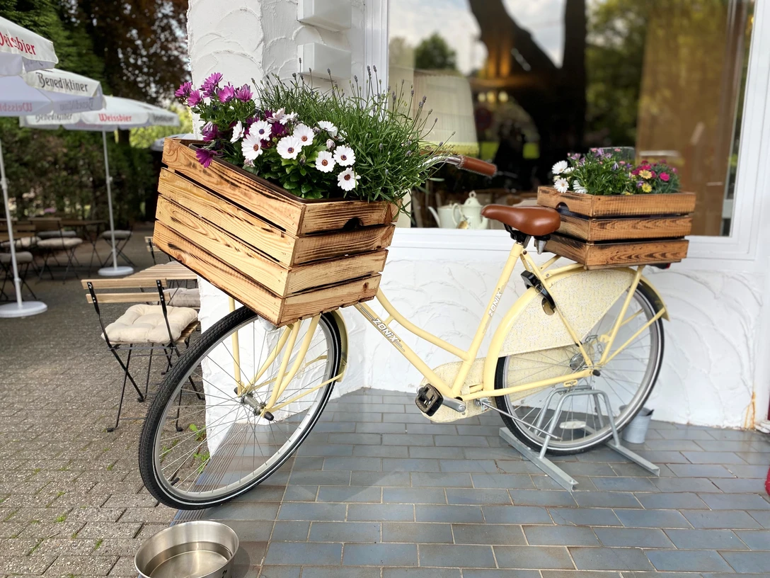 Restaurant Wohnzimmer Neviges Ein vintage gelbes Fahrrad, dekoriert mit blühenden Holzblumenkästen, lehnt an einer weißen Wand.