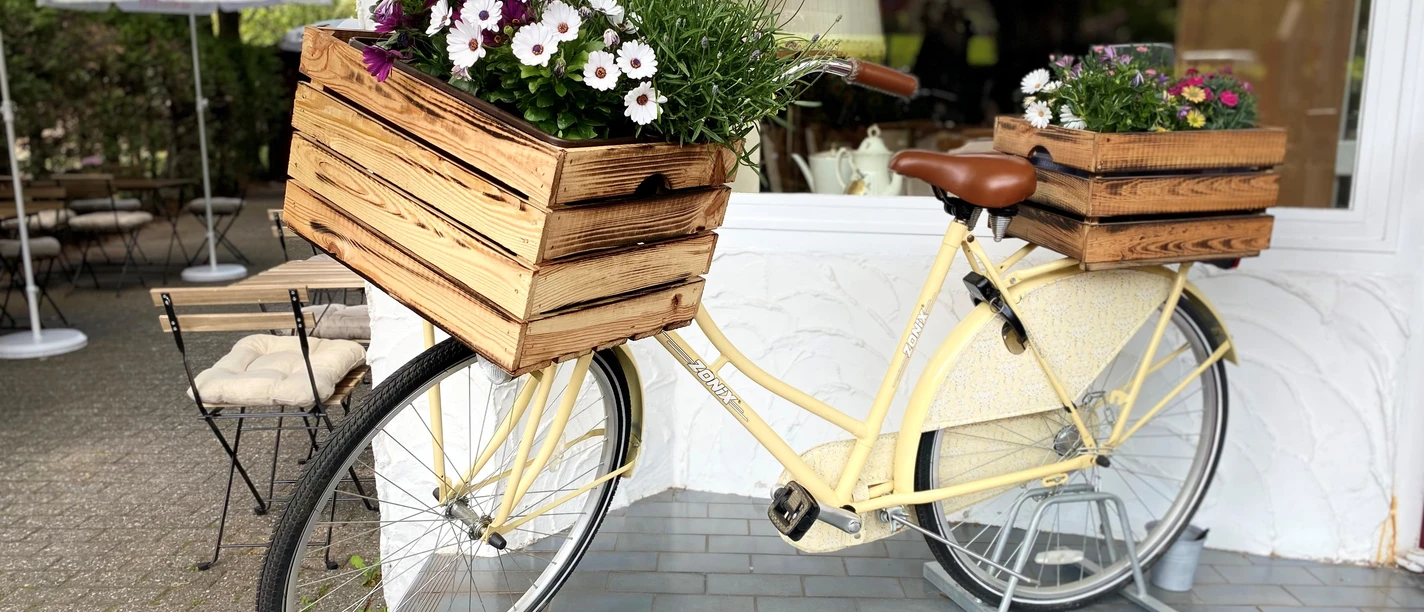 Restaurant Wohnzimmer Neviges Ein vintage gelbes Fahrrad, dekoriert mit blühenden Holzblumenkästen, lehnt an einer weißen Wand.