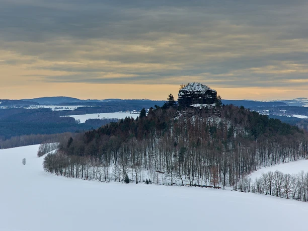 Snow-covered hills and barren trees in front of a rocky cliff, surrounded by vast plains in winter.