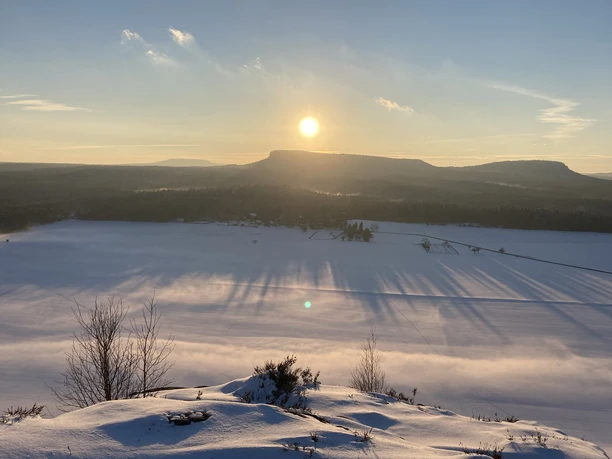 Besneeuwd landschap bij zonsondergang, met lange schaduwen op een bevroren meer en een heuvel op de achtergrond.