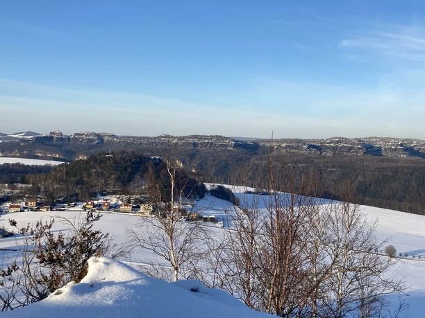 Zasněžená krajina s kopci v pozadí, modrá obloha, stromy v popředí a vesnice s barevnými domy.