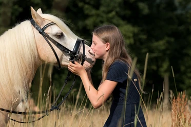 Familien-Reitschule in Velbert Ein Mädchen mit langen, blonden Haaren gibt einem weißen Pferd mit Zaumzeug in einer Wiese einen Kuss.
