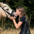 Familien-Reitschule in Velbert Ein Mädchen mit langen, blonden Haaren gibt einem weißen Pferd mit Zaumzeug in einer Wiese einen Kuss.