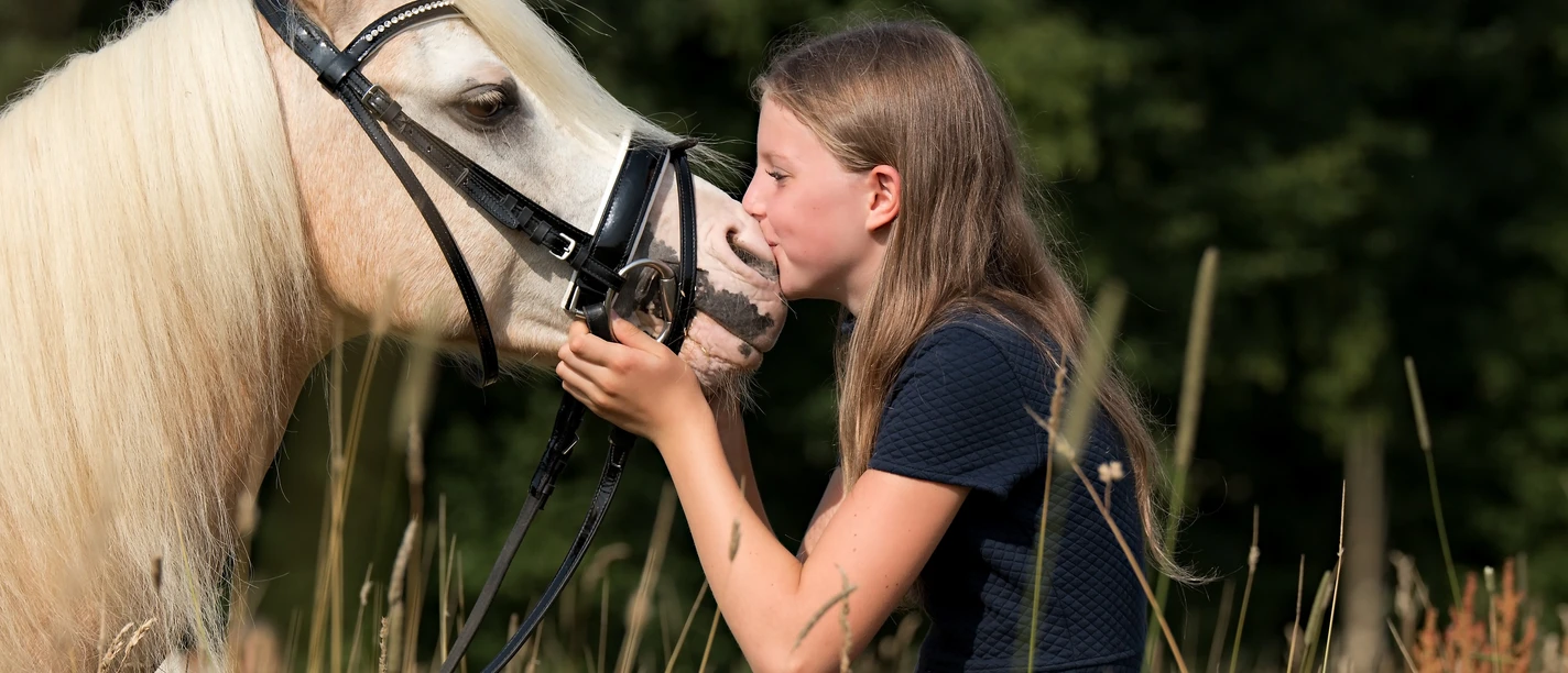 Familien-Reitschule in Velbert Ein Mädchen mit langen, blonden Haaren gibt einem weißen Pferd mit Zaumzeug in einer Wiese einen Kuss.