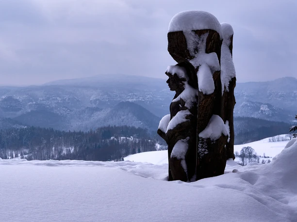 Snow-covered wooden sculpture in front of a wintry mountain landscape with wooded hills and a cloudy sky.