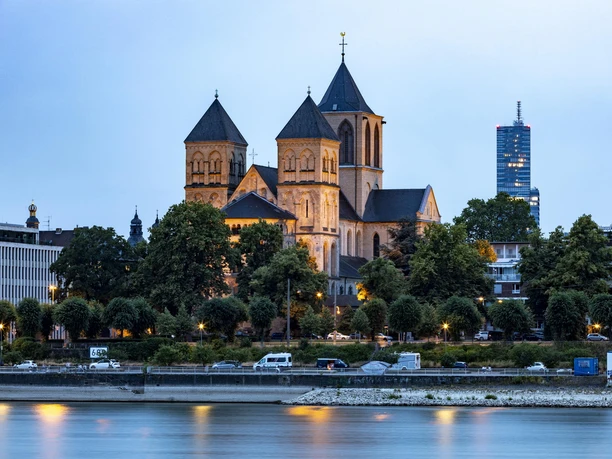 Sankt Kunibert Die romanische Kirche Sankt Kunibert in Köln erhebt sich mit ihren markanten Türmen über den Rhein, flankiert von moderner Architektur im Hintergrund.
