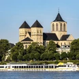 Saint Kunibert Ein romanisches Kirchenbauwerk mit drei Türmen hebt sich majestätisch vor einem klaren Himmel ab. Davor gleitet ein elegantes Ausflugsboot sanft entlang des Rheins, während grüne Bäume das Ufer säumen.A Romanesque church with three towers stands out majestically against a clear sky. In front of it, an elegant excursion boat glides gently along the Rhine, while green trees line the banks.