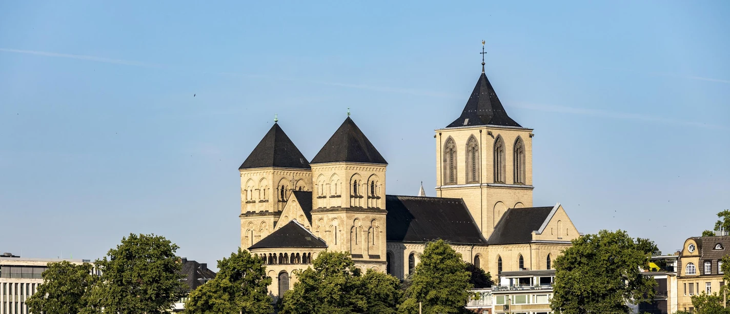 A Romanesque church with three towers stands out majestically against a clear sky. In front of it, an elegant excursion boat glides gently along the Rhine, while green trees line the banks.