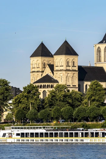Saint Kunibert Ein romanisches Kirchenbauwerk mit drei Türmen hebt sich majestätisch vor einem klaren Himmel ab. Davor gleitet ein elegantes Ausflugsboot sanft entlang des Rheins, während grüne Bäume das Ufer säumen.A Romanesque church with three towers stands out majestically against a clear sky. In front of it, an elegant excursion boat glides gently along the Rhine, while green trees line the banks.