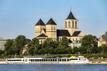 Sankt Kunibert Ein romanisches Kirchenbauwerk mit drei Türmen hebt sich majestätisch vor einem klaren Himmel ab. Davor gleitet ein elegantes Ausflugsboot sanft entlang des Rheins, während grüne Bäume das Ufer säumen.