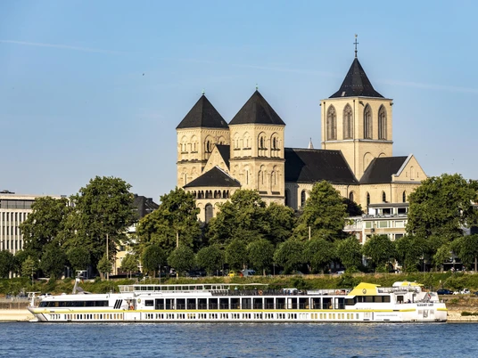 Sankt Kunibert Ein romanisches Kirchenbauwerk mit drei Türmen hebt sich majestätisch vor einem klaren Himmel ab. Davor gleitet ein elegantes Ausflugsboot sanft entlang des Rheins, während grüne Bäume das Ufer säumen.A Romanesque church with three towers stands out majestically against a clear sky. In front of it, an elegant excursion boat glides gently along the Rhine, while green trees line the banks.