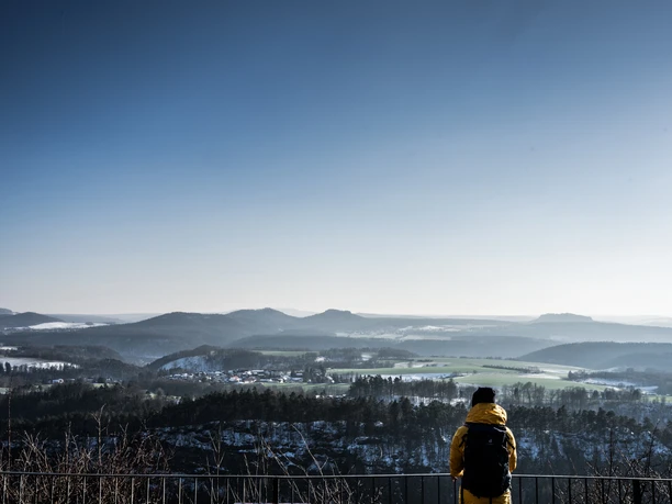 Een persoon in een geel jack met rugzak kijkt vanaf een uitkijkpunt uit over een winters, heuvelachtig landschap onder een strakblauwe hemel.