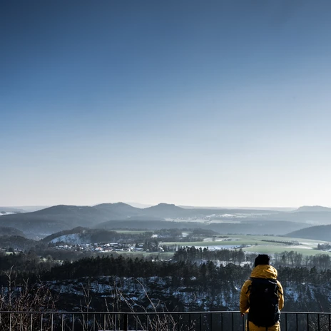 Brandaussicht im Winter Person in gelber Jacke mit Rucksack blickt von einem Aussichtspunkt auf eine winterliche, hügelige Landschaft unter klarem, blauem Himmel.