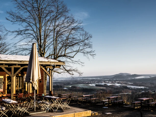 Terrace of a café with empty tables and chairs, snow-covered roof and parasol, in the background a wintry landscape under a blue sky.
