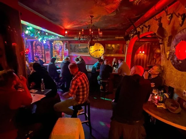 Trash Eine atmosphärische Bar mit roten Lichtern, Wanduhr und Gästen auf Hockern im Gespräch versunken.