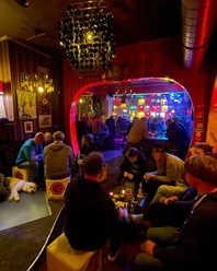 Trash Eine lebhafte Bar mit bunt beleuchtetem Ambiente, Gästen an Tischen und einem Hund im Vordergrund.