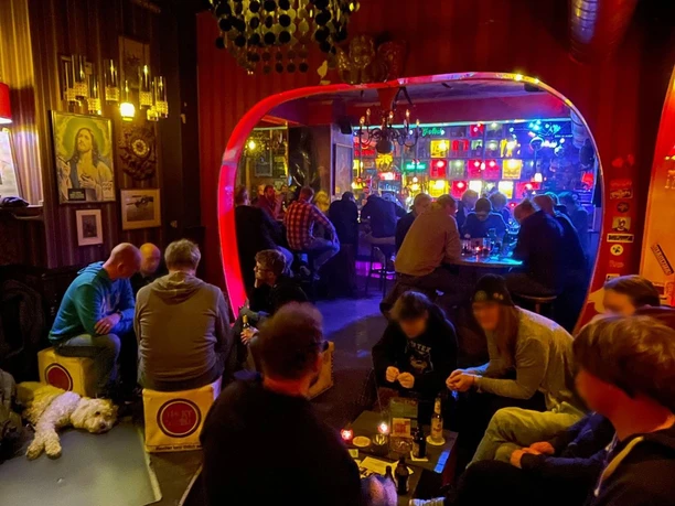 Trash Eine lebhafte Bar mit bunt beleuchtetem Ambiente, Gästen an Tischen und einem Hund im Vordergrund.
