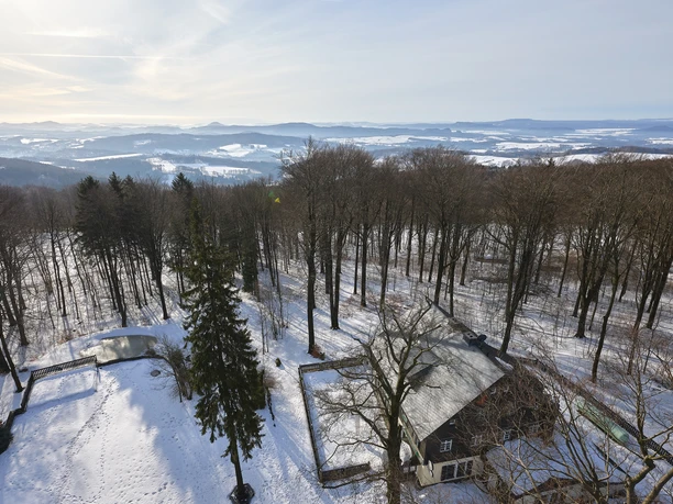 Aussicht vom Ungerturm im Winter Verschneite Landschaft mit einem Haus am Waldrand, umgeben von kahlen Bäumen und einem kleinen Teich, unter einem klaren Himmel.