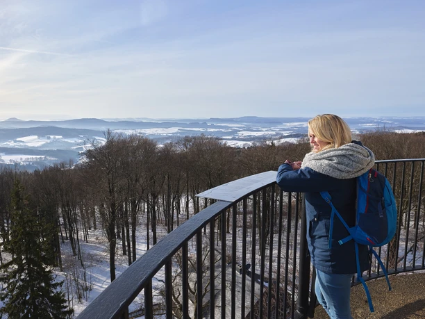 A woman with a blue rucksack stands on a vantage point and looks out over a snow-covered landscape of forests and hills.