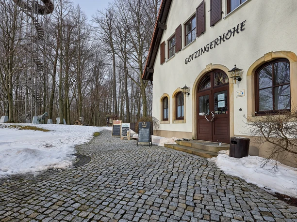 Vchod do budovy s nápisem "Gotzingerhöhe" vedle zasněžené cesty; v pozadí rozhledna a stromy.