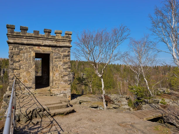 Stone lookout tower with battlements on a rock, surrounded by bare trees and a clear blue sky in the background.