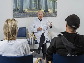 DSCF5603 Gesprächssituation final.jpg Ein Arzt im weißen Kittel spricht engagiert mit zwei Patienten in einem Gesprächsraum.