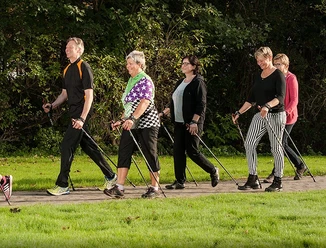 BK_Walking_B1000px - zugeschnitten.jpg Fünf Erwachsene gehen auf einem ebenen Weg durch eine grüne, parkähnliche Umgebung mit Nordic-Walking-Stöcken.