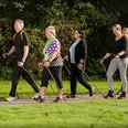 BK_Walking_B1000px - zugeschnitten.jpg Fünf Erwachsene gehen auf einem ebenen Weg durch eine grüne, parkähnliche Umgebung mit Nordic-Walking-Stöcken.