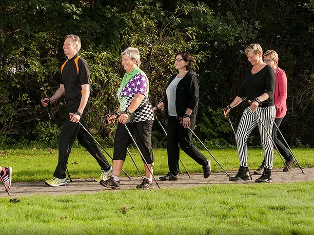 BK_Walking_B1000px - zugeschnitten.jpg Fünf Erwachsene gehen auf einem ebenen Weg durch eine grüne, parkähnliche Umgebung mit Nordic-Walking-Stöcken.