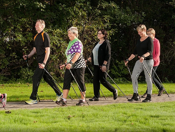 BK_Walking_B1000px - zugeschnitten.jpg Fünf Erwachsene gehen auf einem ebenen Weg durch eine grüne, parkähnliche Umgebung mit Nordic-Walking-Stöcken.