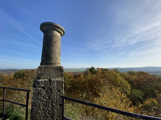 Triangulation Column Cotta Spitzberg Stone pillar on a vantage point, surrounded by autumnal trees and a sweeping view of the hilly landscape under a blue sky.